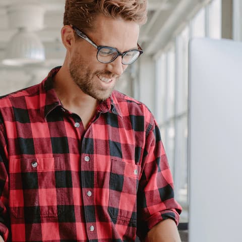 Standing and smiling light-skinned man with beard stubble wearing plaid shirt and glasses